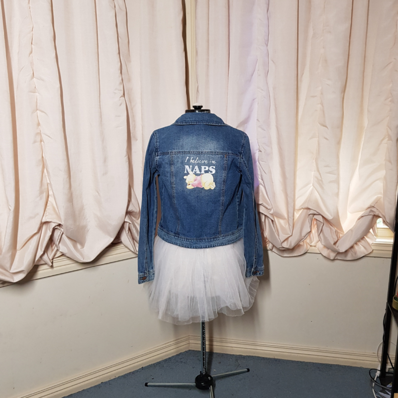 A "I Believe in Naps" Upcycled Denim Jacket displayed on a mannequin in front of curtains.